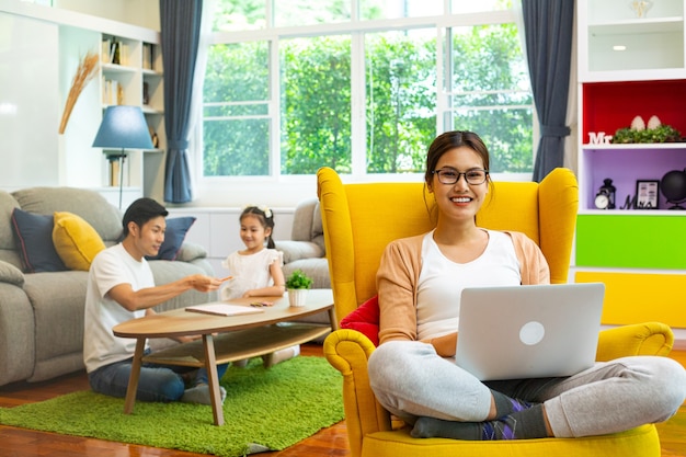 La mamma si siede e lavora a casa sul divano giallo. Famiglia asiatica che fa attività insieme.