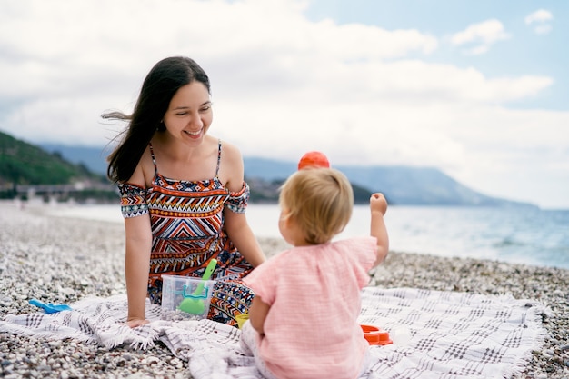 La mamma sorride alla bambina con uno stampo giocattolo sulla testa mentre è seduta su una spiaggia di ciottoli
