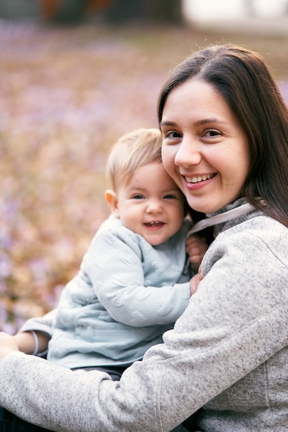 La mamma sorridente abbraccia il bambino sorridente mentre è seduto sul fogliame nel ritratto del primo piano del parco