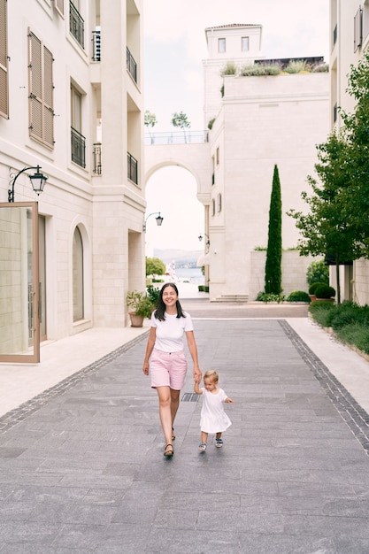 La mamma sorridente conduce una bambina per mano lungo una strada acciottolata tra grattacieli