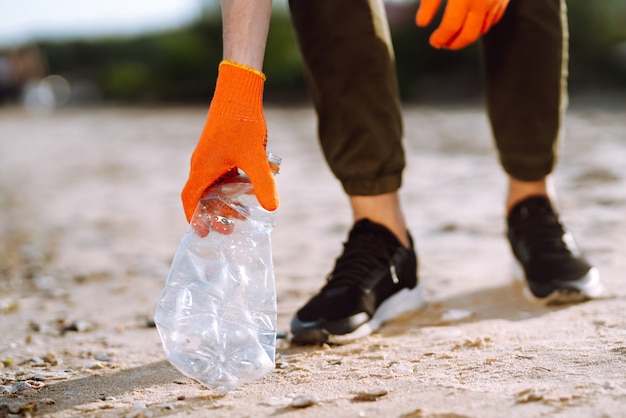 La mano degli uomini raccoglie la bottiglia di plastica sulla spiaggia del mare. Il volontario che indossa guanti protettivi raccoglie la bottiglia di plastica.
