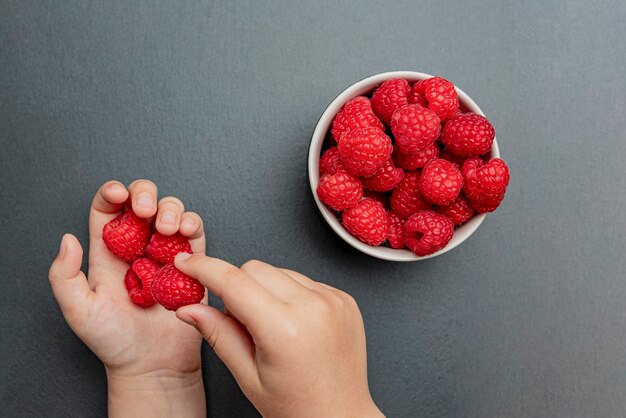 La mano del bambino prende lamponi freschi e succosi da un piccolo piatto nero. Primo piano cremisi rosso brillante. Tempo di raccolta delle bacche estive. Frutti biologici sani per i bambini.