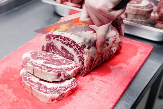 La mano del cuoco del primo piano taglia la bistecca di manzo marmorizzata ribeye con il coltello tagliente sul tagliere di plastica rosso sulla tavola del metallo