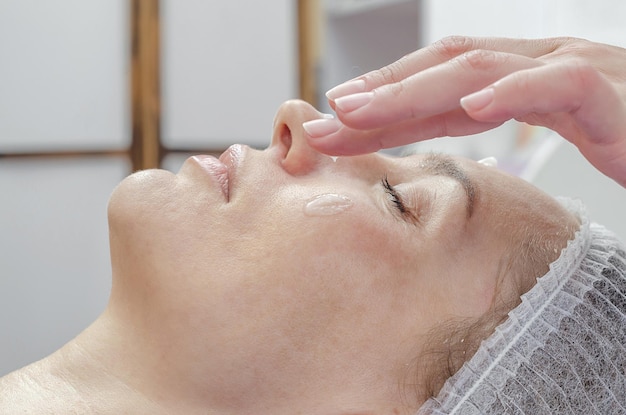 La mano dell'estetista applica la crema sul viso della donna nel salone spa Viso rilassato sul divano Pelle ringiovanente