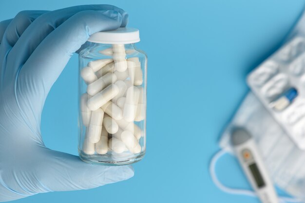 La mano dell'infermiera in un guanto medico blu detiene una lattina di vetro con pillole su sfondo blu. Blister protettivo della maschera, del termometro e delle pillole della medicina defocused su fondo.