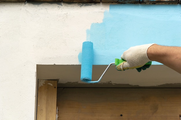 La mano dell'uomo dipinge un muro sopra una finestra o una porta con vernice blu.
