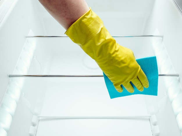 La mano dell'uomo nei guanti che pulisce il frigorifero bianco con uno straccio blu in cucina