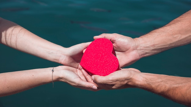 La mano della coppia che tiene la forma rossa del cuore contro il fondo dell&#39;acqua di mare