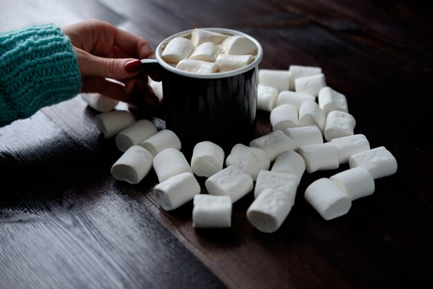 La mano della donna che tiene tazza di caffè nera con le caramelle gommosa e molle fra le caramelle gommosa e molle su superficie scura di legno