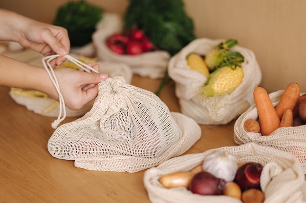 La mano della donna chiude una borsa della spesa riutilizzabile con verdure su un tavolo a casa in cotone a rete con