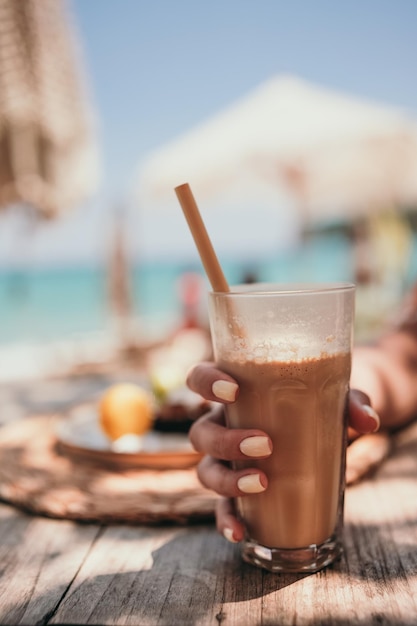 La mano della donna con la manicure gialla tiene il latte freddo del caffè in vetro alto con paglia in riva al mare nel bar sulla spiaggia