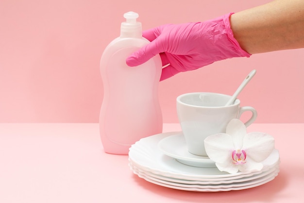 La mano della donna in un guanto protettivo con una bottiglia di detersivo per piatti piatti bianchi un piattino una tazza