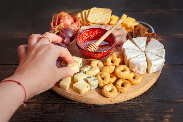 La mano della donna prende un pezzo di formaggio dalla tavola degli antipasti con formaggio assortito carne salsiccia rosetta uva e biscotti tagliere di salumi e formaggi Vista dall'alto