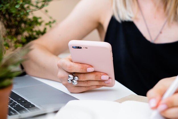 La mano della donna tiene lo smartphone e prende appunti nel blocco note nell'accogliente ufficio estivo con il computer portatile