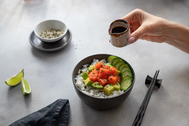 La mano della donna tiene una salsiera con salsa di soia sopra una ciotola scura con salmone, riso, avocado e cetriolo. insalata tradizionale di pesce crudo hawaiano.