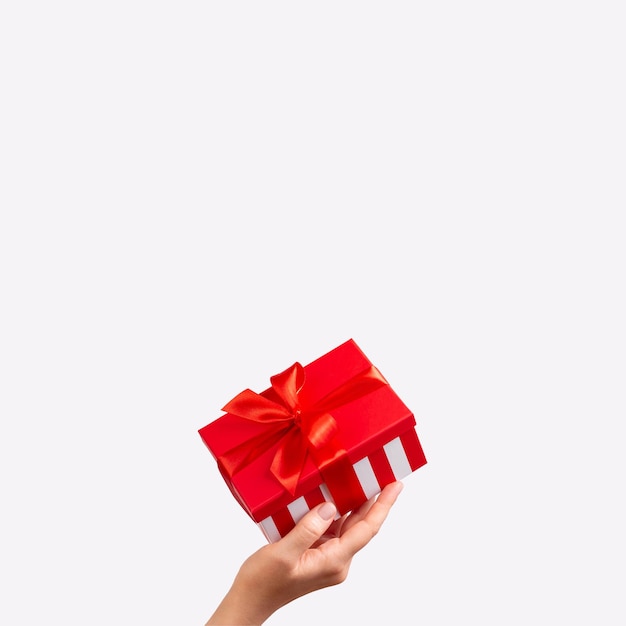 La mano della donna tiene una scatola regalo a righe con nastro rosso su sfondo bianco Natale Capodanno San Valentino e compleanno spazio di copia del concetto