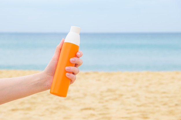 La mano della donna tiene uno spray solare in una bottiglia arancione sullo sfondo di una spiaggia sabbiosa e di un mare