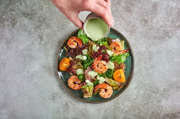 La mano della donna versa la salsa su insalata con gamberi fritti, pomodori gialli, rucola, spinaci, lattuga e salsa su una piastra in ceramica