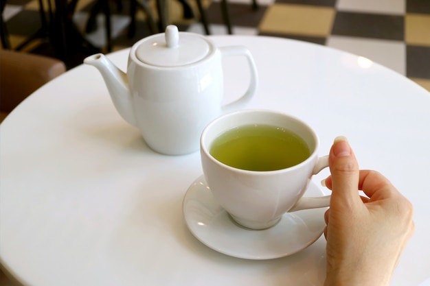 La mano della femmina che tiene tazza di tè verde su una tavola rotonda bianca con la teiera nel fondo