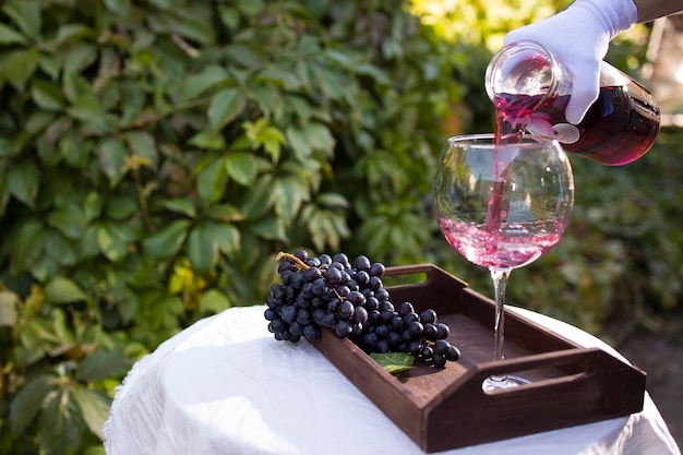 La mano di un cameriere in un guanto bianco versa un bicchiere di vino rosso che si trova su un tavolo con una tovaglia bianca all'aperto alla luce del sole