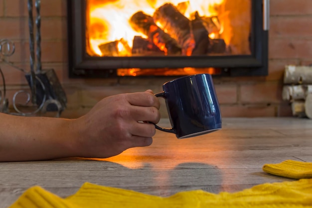 La mano di un uomo tiene una tazza con una bevanda calda vicino al camino con un fuoco Tenere al caldo in una casa calda in inverno