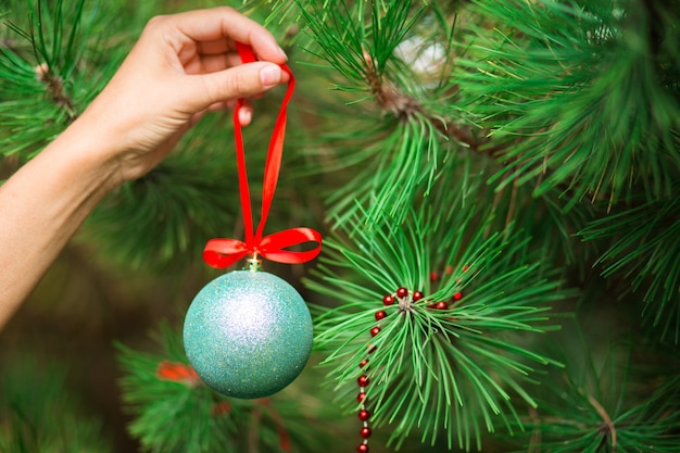 La mano di una donna appende una palla di Natale con un nastro rosso su un ramo di pino