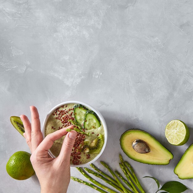 La mano di una donna mette un piatto di asparagi su uno sfondo di cemento grigio con un frullato di dieta verde