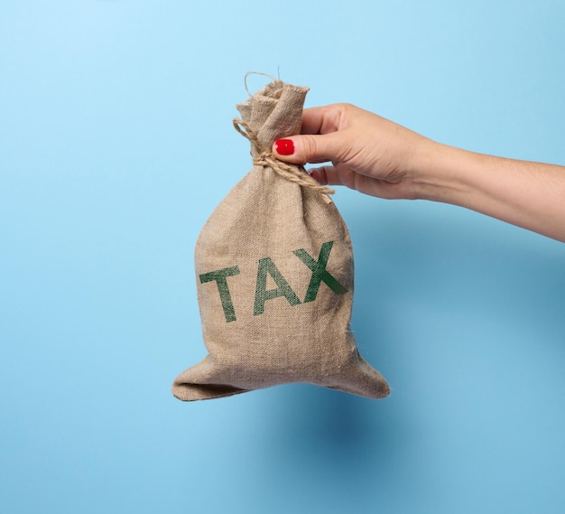 La mano di una donna tiene in mano un sacco di iuta pieno con la tassa di iscrizione su sfondo blu che rappresenta il concetto di aumento delle tasse