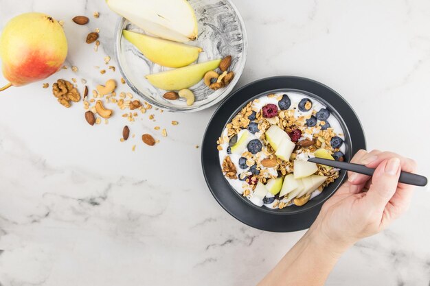 La mano di una giovane donna con un cucchiaio di granola riempito durante la colazione. piatto con muesli, frutti di bosco, pera matura. cibo misto, fitness, dieta.