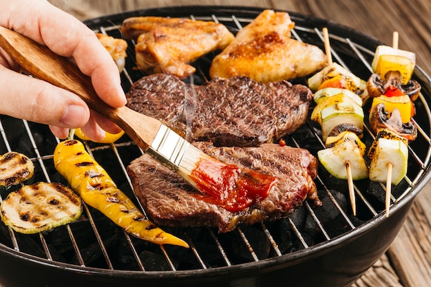 La mano di una persona che spande la salsa sulla bistecca arrostita sopra il barbecue