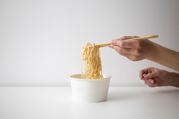 La mano femminile prende le tagliatelle cinesi con le bacchette cinesi da una ciotola di carta su una priorità bassa bianca