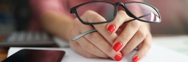 La mano femminile tiene gli occhiali accanto a smartphone e penna.