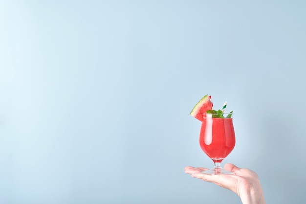 La mano femminile tiene il cocktail di anguria in vetro con menta e ghiaccio su sfondo blu. Vacanze estive calde colorate. Chiudere le fette di anguria nelle mani. Bandiera. Modello.