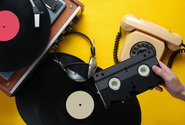 La mano femminile tiene la videocassetta. Stile anni '80. Lettore vinile, cuffie, telefono rotativo su sfondo giallo. Vista dall'alto, piatto