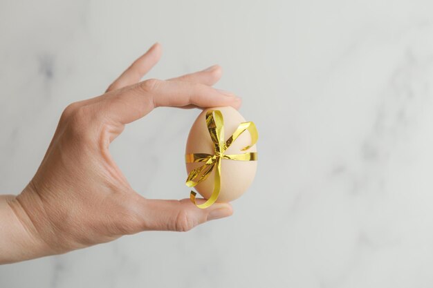 La mano femminile tiene un grande uovo di gallina legato con un nastro festivo su sfondo grigio astratto