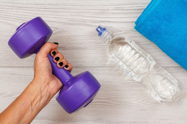 La mano femminile tiene un manubrio in camera o in palestra con una bottiglia d'acqua e un asciugamano sullo sfondo. Strumenti per il fitness. Vista dall'alto