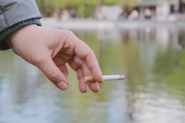 La mano femminile tiene una sigaretta accesa sullo sfondo di uno stagno in un parco cittadino. Tendenza a fumare. Affrontare lo stress emotivo con il fumo. Spazio per il testo.