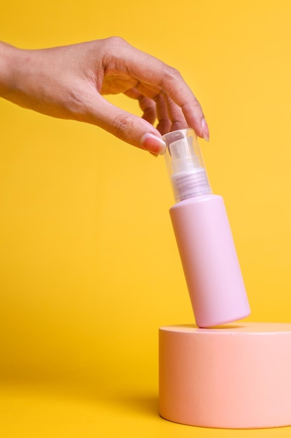 La mano femminile tocca un flacone spray rosa di profumo per il corpo su un podio rosa isolato su sfondo giallo