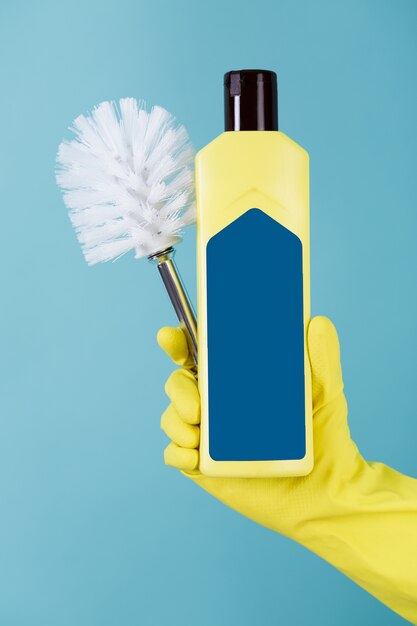 La mano in guanto giallo tiene la bottiglia di detersivo liquido per la toilette e la spazzola su fondo blu.