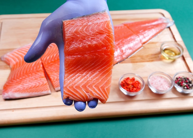 La mano maschile in guanti blu tiene un pezzo fresco di filetto di salmone crudo e sullo sfondo salmone e spezie su un tagliere di legno wooden