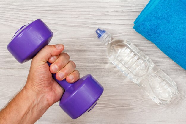 La mano maschile tiene in mano un manubrio in camera o in palestra con una bottiglia d'acqua e un asciugamano sullo sfondo. Strumenti per il fitness. Vista dall'alto