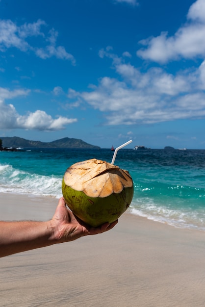 La mano maschio sta tenendo i cocktail freschi di una noce di cocco della noce di cocco sulla bella spiaggia a Sunny Day.