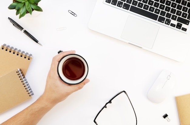 La mano maschio tiene una tazza di caffè vicino a laptop, occhiali e accessori