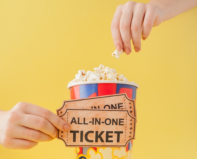 La mano prende biglietti per il cinema e popcorn da un bicchiere di carta su uno sfondo giallo. La donna mangia popcorn. Concetto di cinema. Disteso.