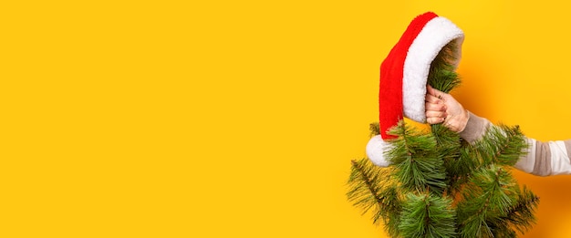 La mano tiene l'albero di Natale con il cappello di Babbo Natale su sfondo giallo. Concetto per Capodanno e vigilia di Natale. Bandiera.