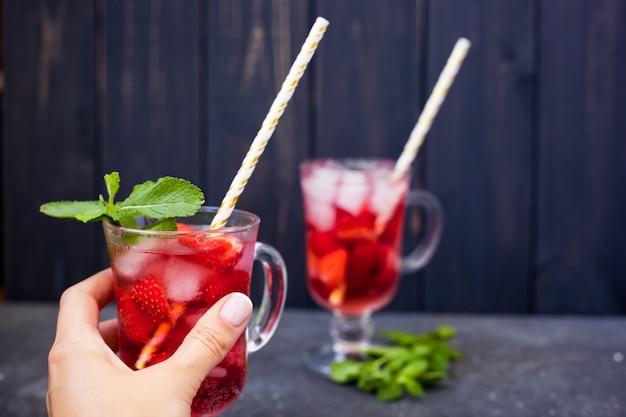 La mano tiene la bevanda della fragola con ghiaccio e le foglie di menta su un fondo scuro