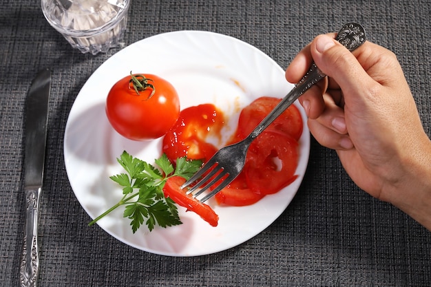 La mano tiene la forchetta che mangia pomodori freschi con un piatto bianco di ketchup su una tovaglia scura