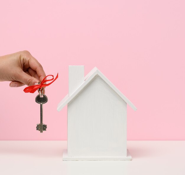 La mano tiene le chiavi e la casa in legno su uno sfondo rosa. Affitto immobiliare, acquisto e concetto di vendita. Servizi di agente immobiliare, riparazione e manutenzione di edifici, spazio di copia