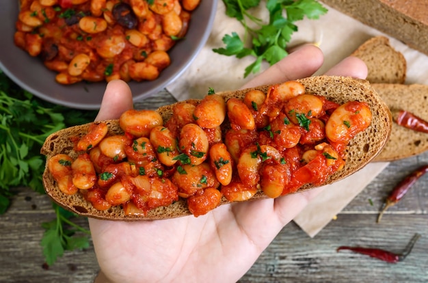 La mano tiene un pezzo di pane di segale con fagioli sullo sfondo del tavolo. Fagioli in umido in salsa di pomodoro con erbe e spezie. T