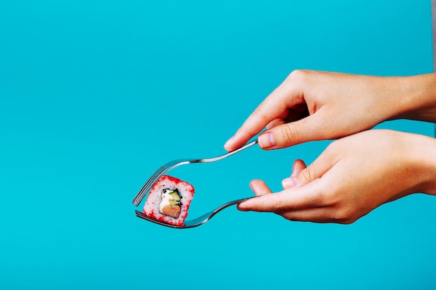 La mano tiene un rotolo di sushi fresco con una forchetta, isolato su sfondo blu. Come mangiare il sushi senza bacchette.